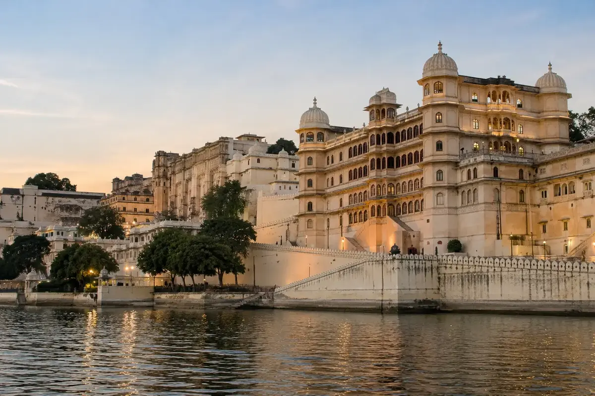 Scenic view of City Palace Udaipur illuminated at dusk, showcasing its grand white marble architecture along the shores of Lake Pichola in Rajasthan.